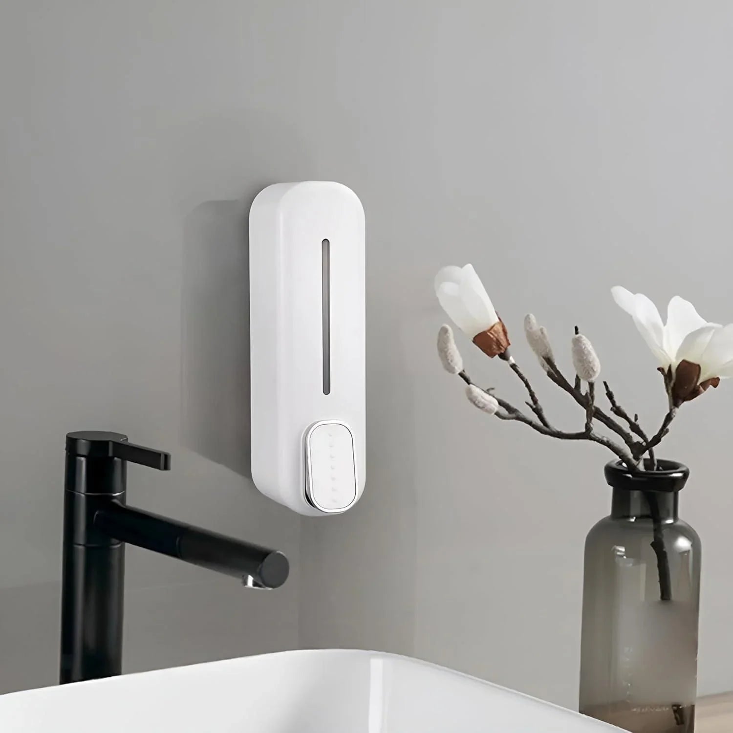 White wall mounted soap dispenser installed above sink next to black faucet and decorative flower vase