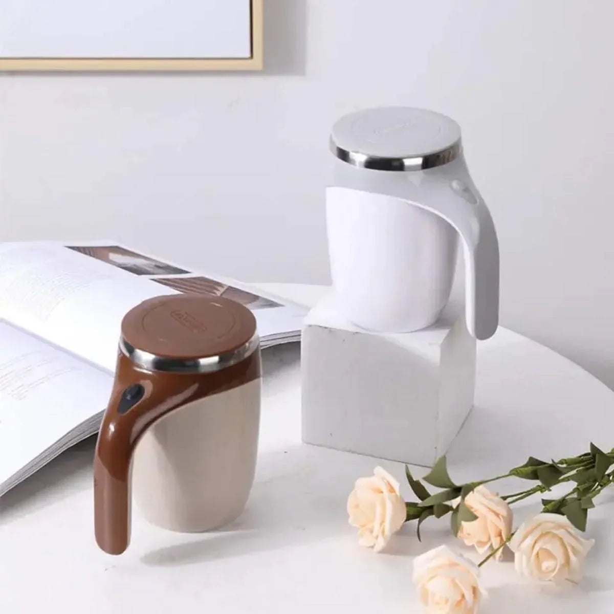 Two modern self stirring mugs in white and beige with brown and white handles on a white table with flowers and a book